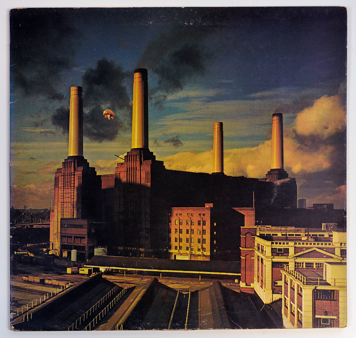 Album cover of Pink Floyd's "Animals" featuring a photograph of Battersea Power Station in London under a cloudy sky. An inflatable pig floats between two of the building's towering chimneys, symbolizing themes of power, greed, and societal division. The industrial structure stands stark against a dark and dramatic sky, evoking a sense of foreboding and critique of capitalist excess.